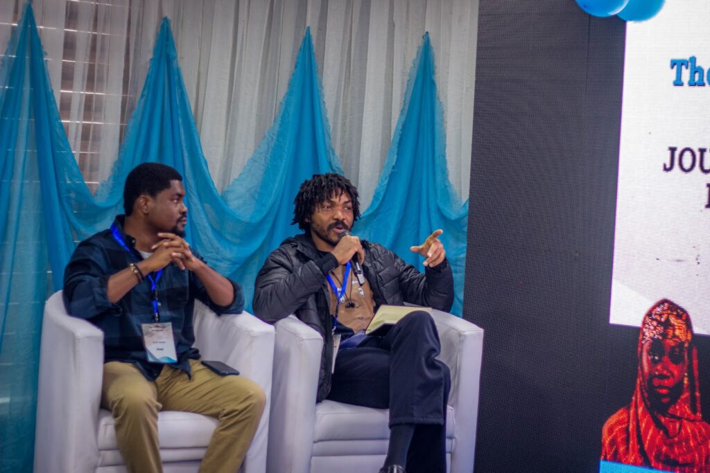 Two men sitting and discussing on a panel stage with blue drapery and screen background.