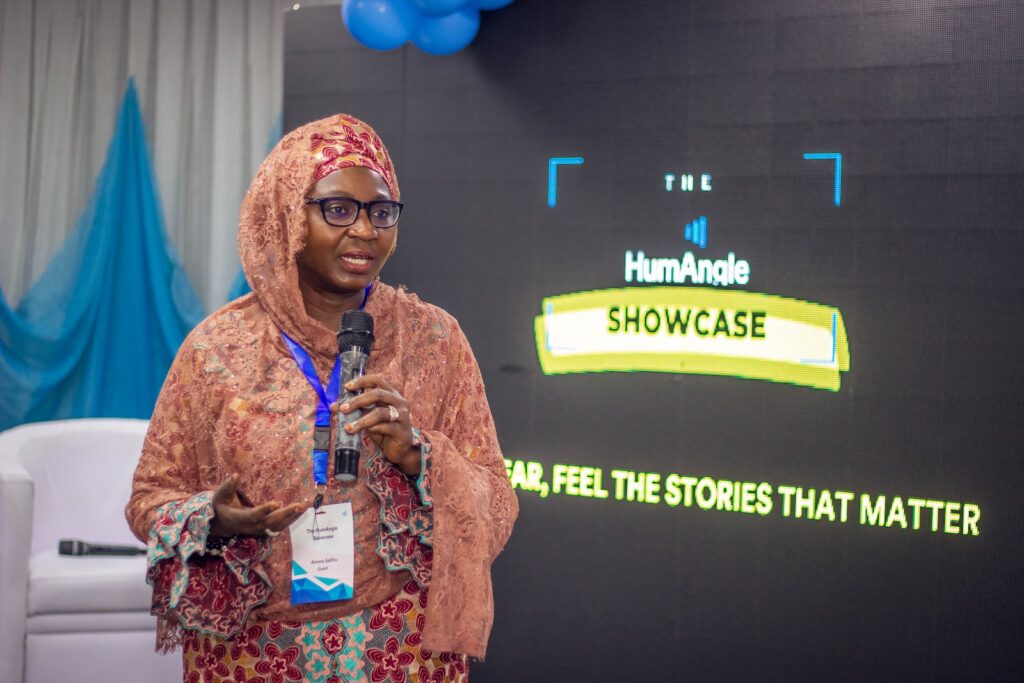 Woman presenting at HumAngle Showcase event, standing with microphone against a digital screen backdrop.