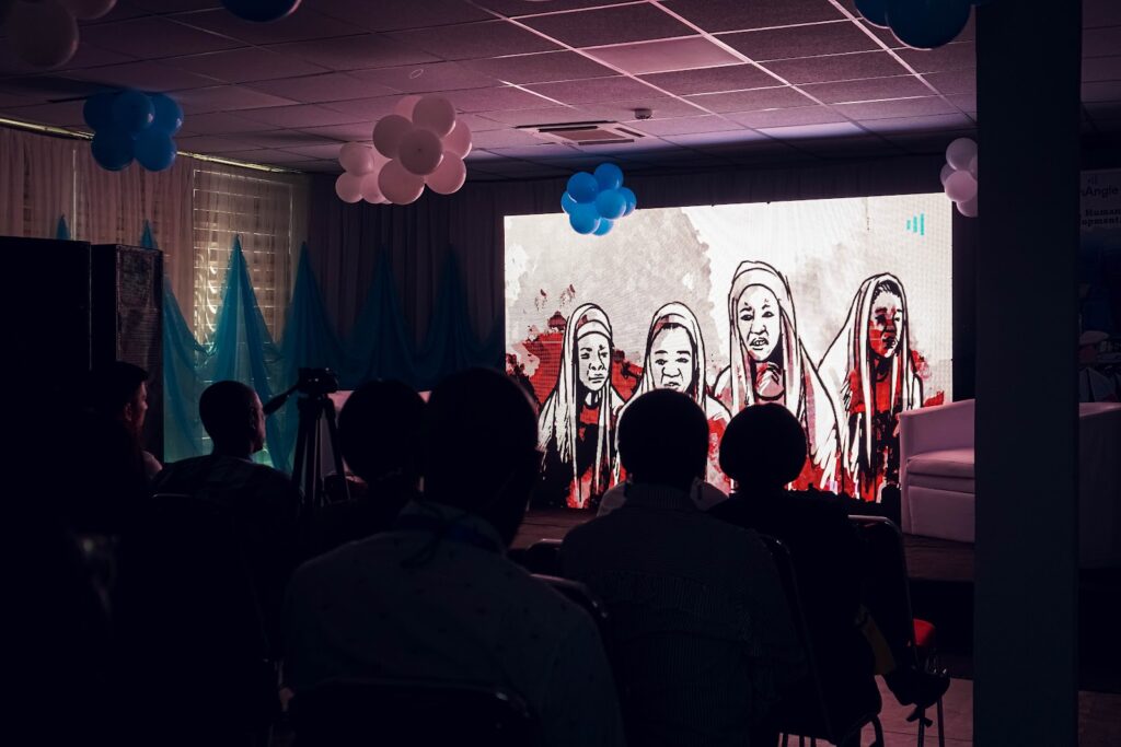 Audience watching a projected art piece featuring four stylized faces at an event with blue and white balloons.