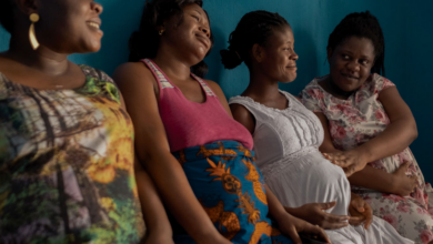 Four women sitting side by side, smiling, with a blue wall in the background.