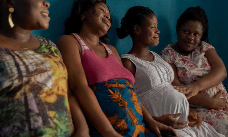 Four women sitting side by side, smiling, with a blue wall in the background.