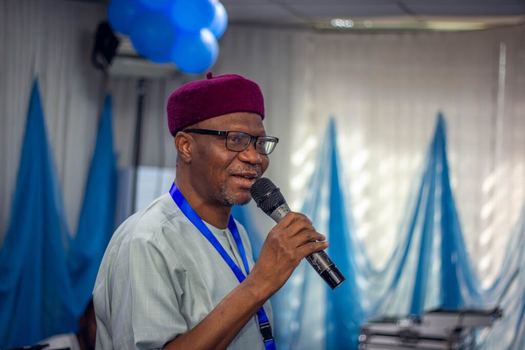 Man in a burgundy hat speaking into a microphone at an event with blue decorations in the background.