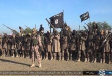 A group of armed individuals in military attire, standing with flags, in a desert location.