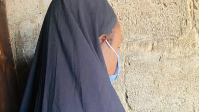 Person in a dark veil leaning against a textured wall, partially facing away.