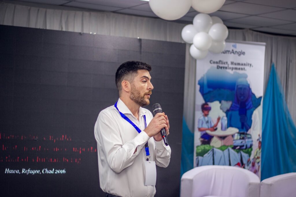 Man speaking into a microphone at an event with a banner in the background.