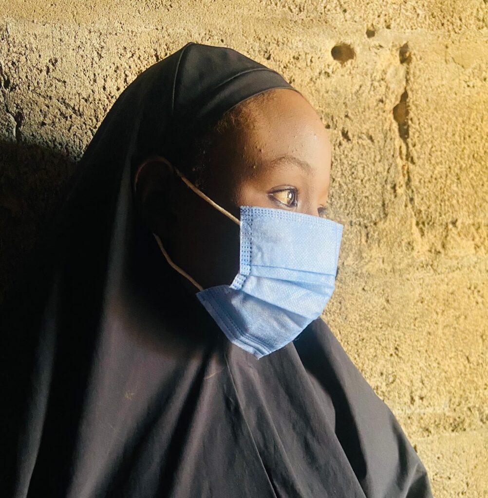 A person in a black hijab and blue face mask looking to the side against a textured wall.