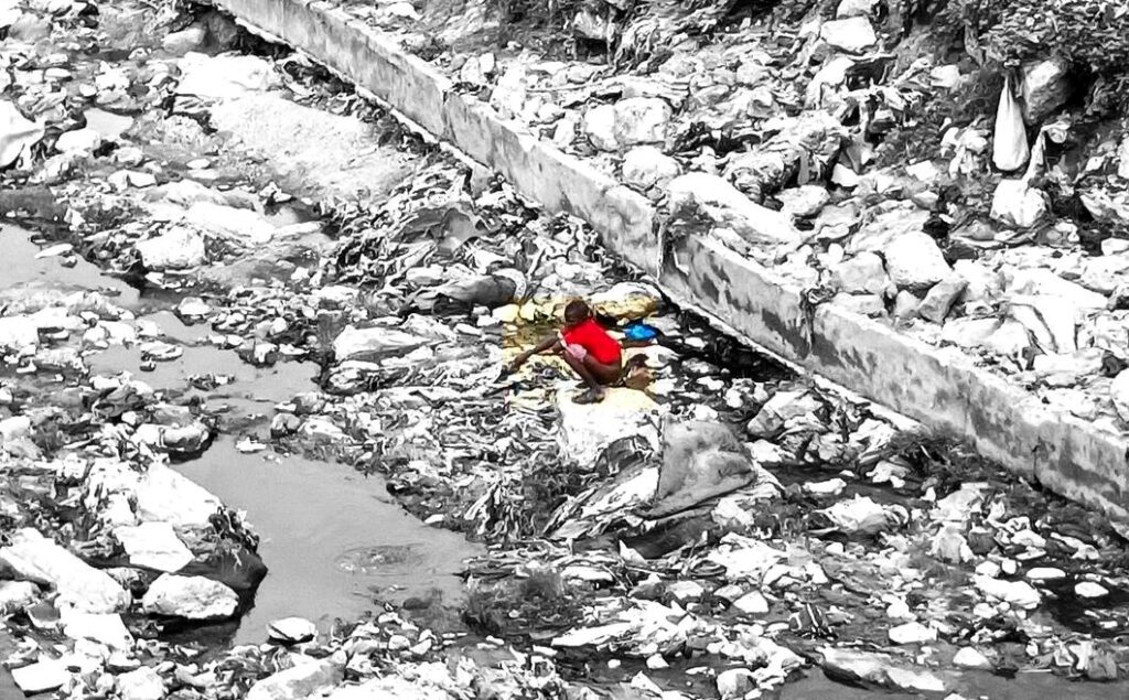 A child in red sitting alone amid a black and white image of a littered streambed, highlighting pollution.