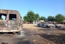 Burnt-out vehicles in a dusty lot with trees and buildings in the background.