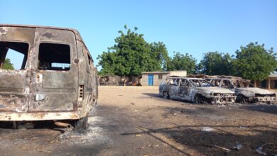 Burnt-out vehicles in a dusty lot with trees and buildings in the background.