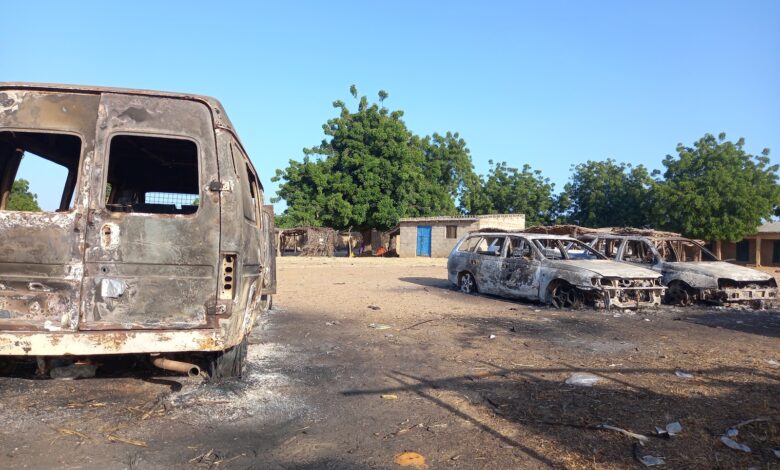 Burnt-out vehicles in a dusty lot with trees and buildings in the background.