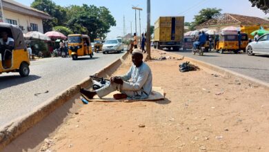 Man sitting on a roadside median with passing traffic and auto rickshaws in a busy street scene.