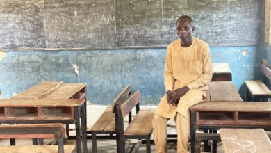 Man seated in a classroom with wooden desks and a chalkboard behind him.