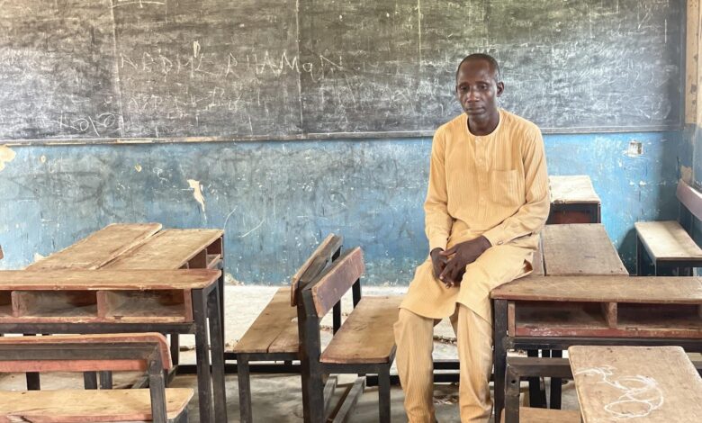 Man seated in a classroom with wooden desks and a chalkboard behind him.