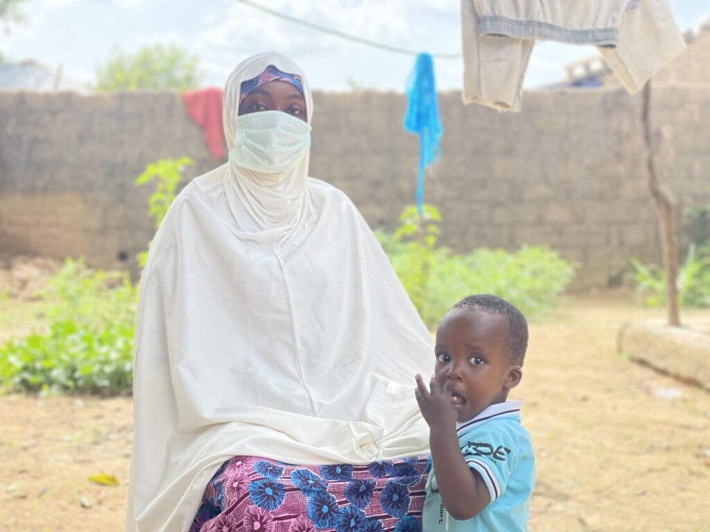 Woman in a white hijab and surgical mask sitting outside with a young child in a blue shirt. Clothes hanging on a line in the background.