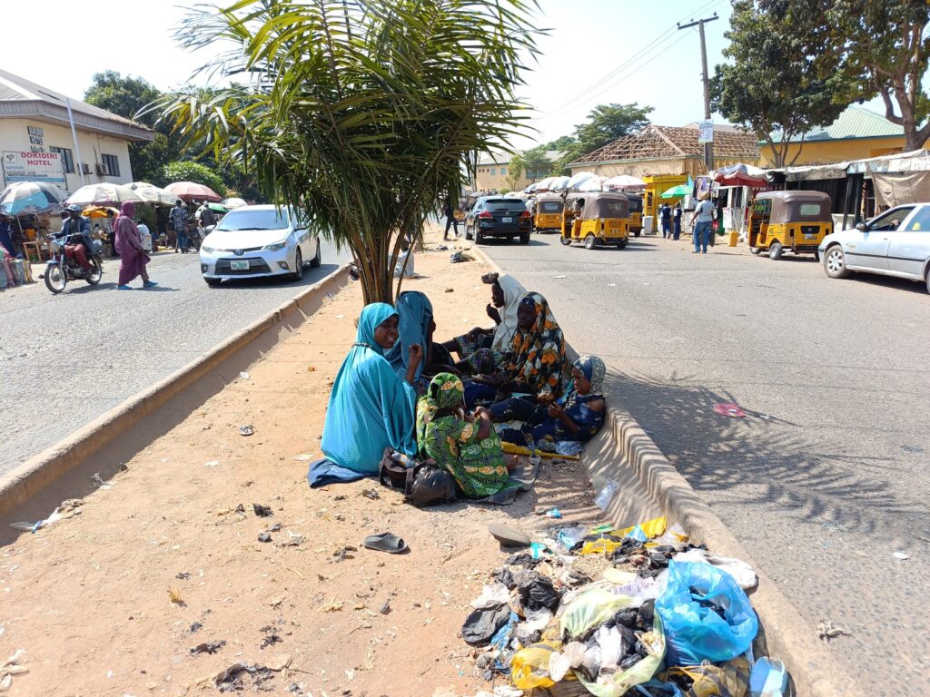 People sitting on the roadside in a busy street market setting with vehicles and trash nearby.