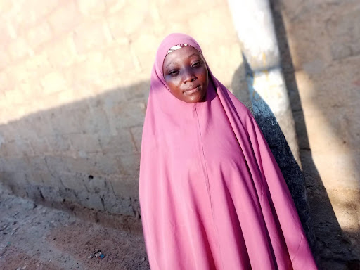 A woman in a pink hijab standing against a wall with a soft smile.