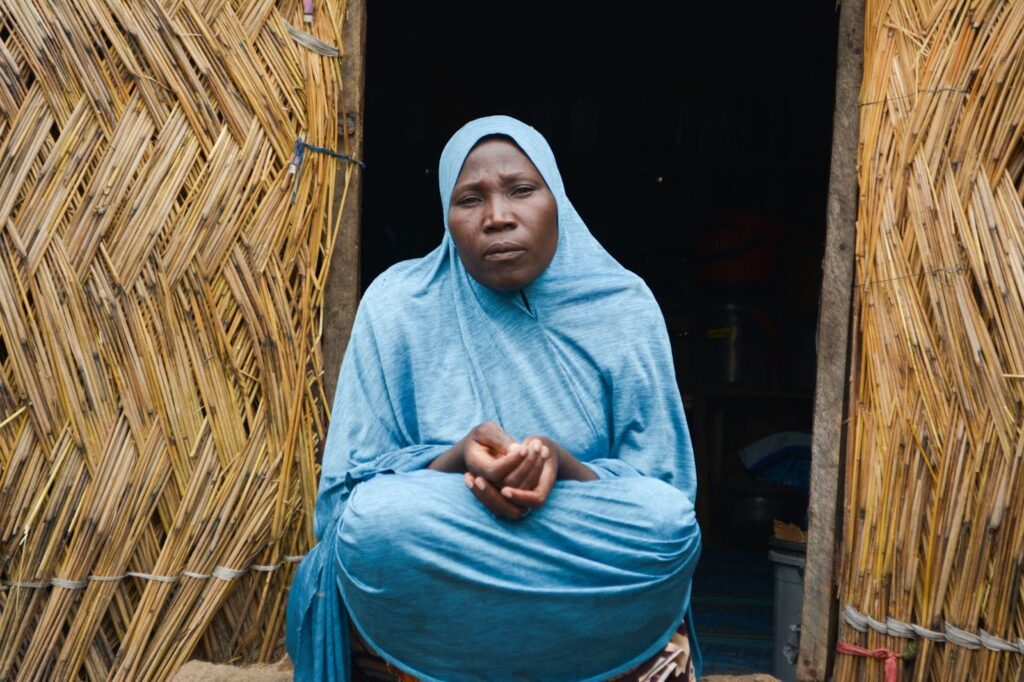 Woman in blue garment sitting at the entrance of a thatched hut.