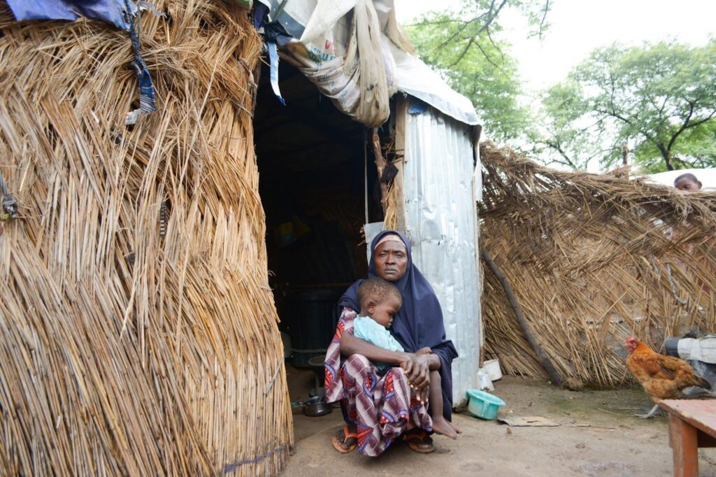 Woman and child sitting by a thatched hut and metal sheet, with a roaming chicken nearby.