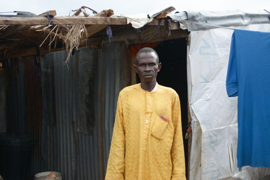 Man in yellow shirt standing before a makeshift shelter with corrugated metal and tarp walls.