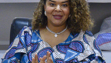 Woman with curly hair in a patterned blouse sits at a desk, gesturing with hands, notebook and pen in front.