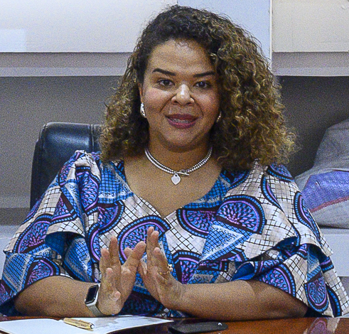 Woman with curly hair in a patterned blouse sits at a desk, gesturing with hands, notebook and pen in front.