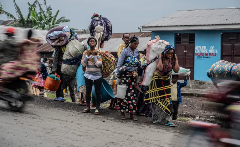 People carrying belongings walk along a road, with a blurred motorcycle passing by.