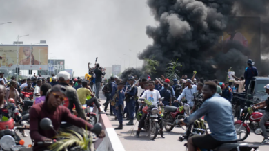 Crowd on motorcycles amidst protest, with people raising hands and black smoke in the background.