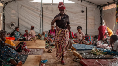 A woman carries a pot in a crowded tent with people sitting on makeshift beds on the ground.