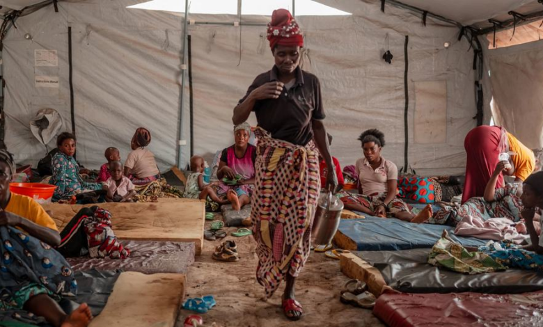 A woman carries a pot in a crowded tent with people sitting on makeshift beds on the ground.