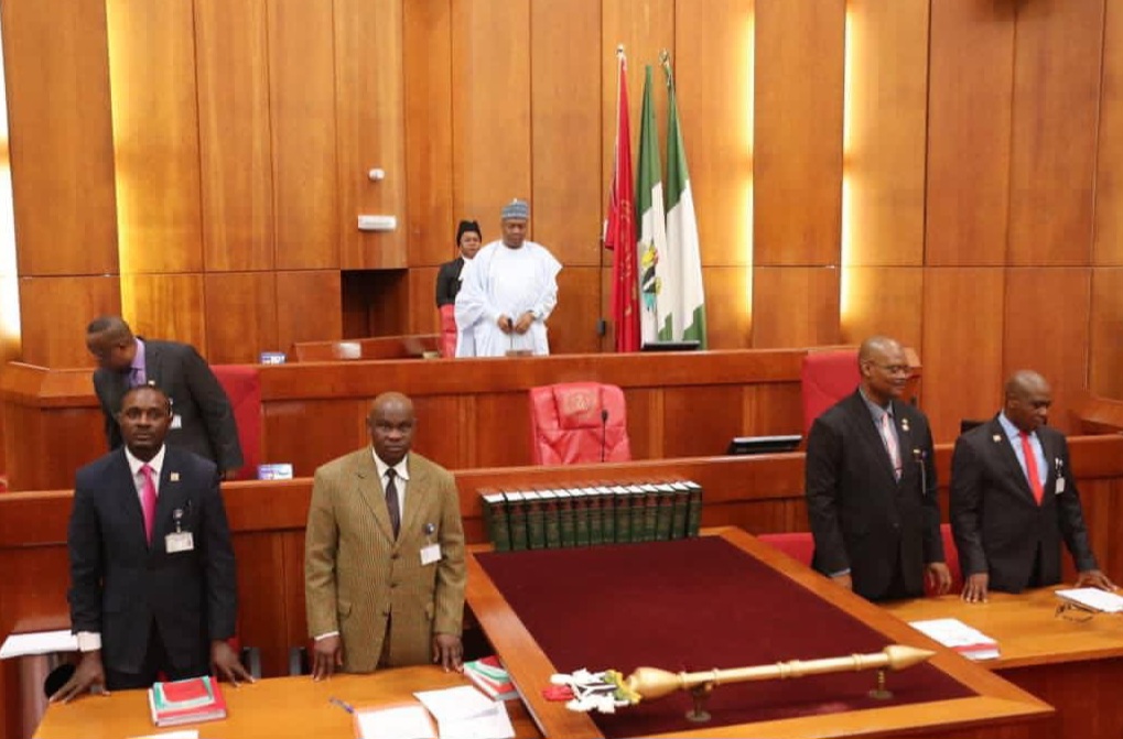 Officials in a governmental chamber, with flags and books visible, prepare for a session. A mace lies on the table in the foreground.