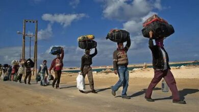 A line of people walking under a blue sky, each carrying large bundles on their heads along a sandy path.