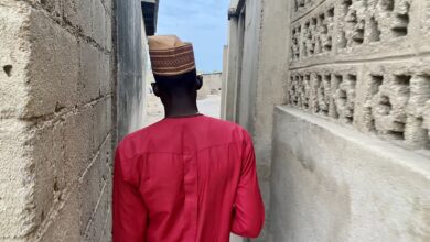 Man in a red shirt and patterned cap walks through a narrow alley bordered by concrete walls.