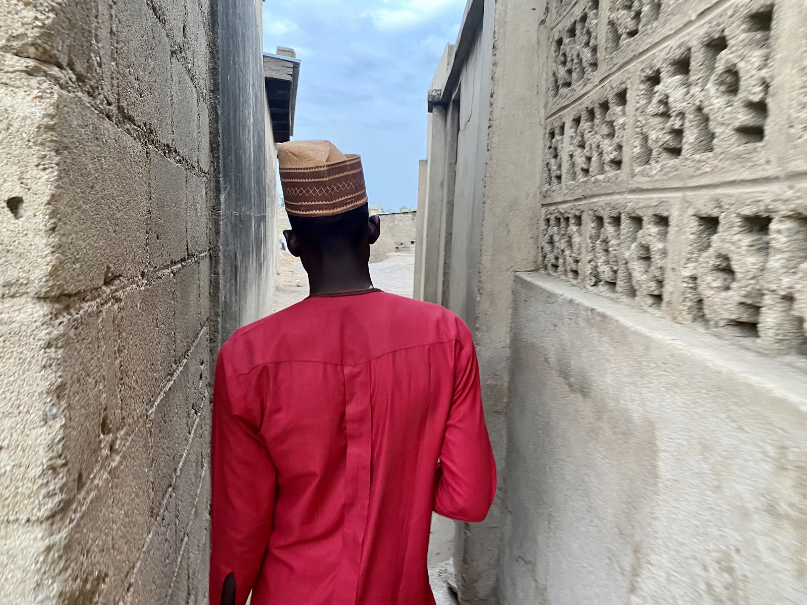 Man in a red shirt and patterned cap walks through a narrow alley bordered by concrete walls.