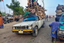 Pickup truck loaded with firewood, carrying passengers on top, drives through a dusty street. People and vehicles are around.