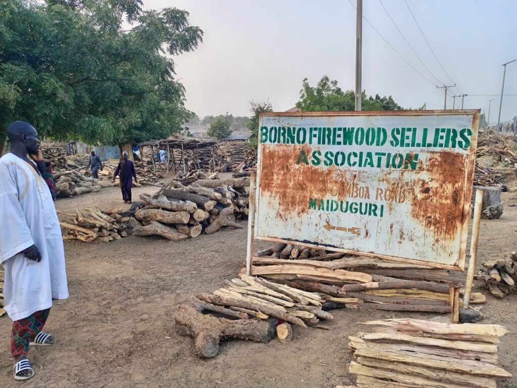 A person stands near a sign for the Borno Firewood Sellers Association in a wood market in Maiduguri. Logs are stacked nearby.