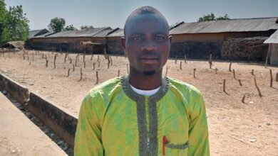 Man in a green patterned shirt standing outdoors on a sunny day, with rustic buildings and trees in the background.