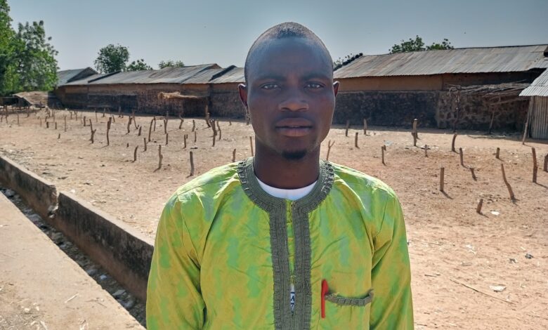 Man in a green patterned shirt standing outdoors on a sunny day, with rustic buildings and trees in the background.