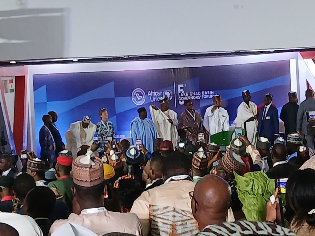 People gathered at the 5th Lake Chad Basin Governors' Forum, African Union leaders on stage, audience taking photos.