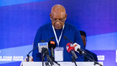 A person in a blue shirt speaks at a podium with multiple microphones against a blue backdrop at a conference.