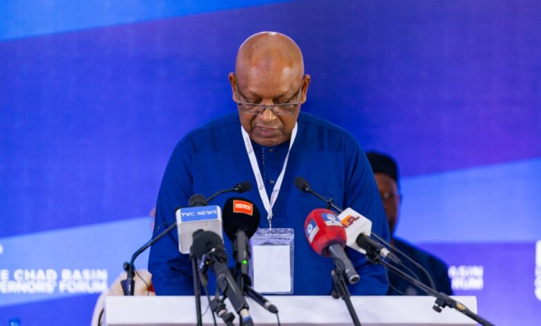 A person in a blue shirt speaks at a podium with multiple microphones against a blue backdrop at a conference.