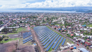 Aerial view of a solar farm with rows of panels near a densely populated town by a body of water.