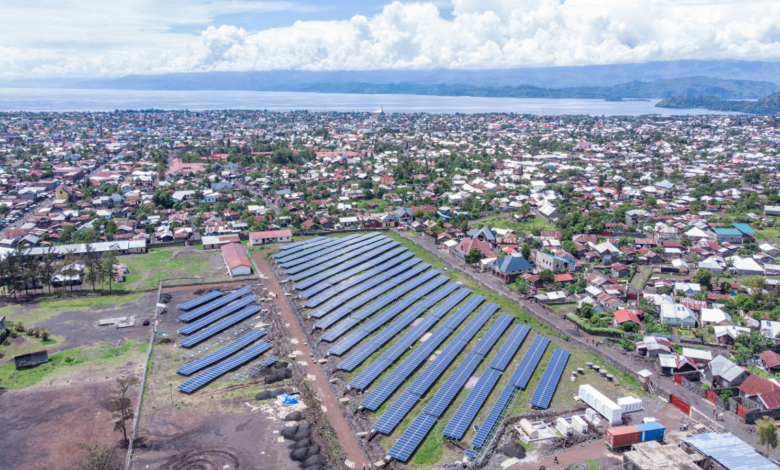 Aerial view of a solar farm with rows of panels near a densely populated town by a body of water.