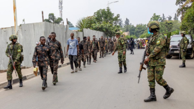 Soldiers escort a group of people along a street, with vehicles and others in the background.