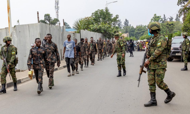 Soldiers escort a group of people along a street, with vehicles and others in the background.