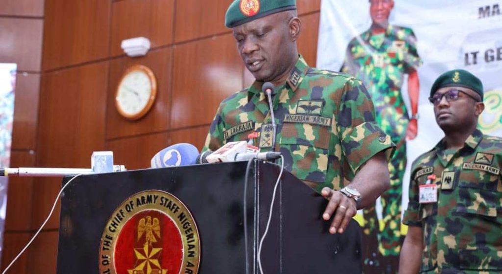 Nigerian Army officer in camouflage speaks at podium during a conference, with another officer standing nearby.