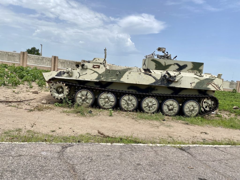 Camouflaged military vehicle parked on a sandy terrain with a partly cloudy sky in the background.