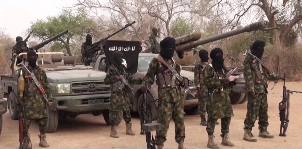 Armed militants in camouflage and masks stand in front of vehicles with mounted weaponry and a black flag in a rural setting.