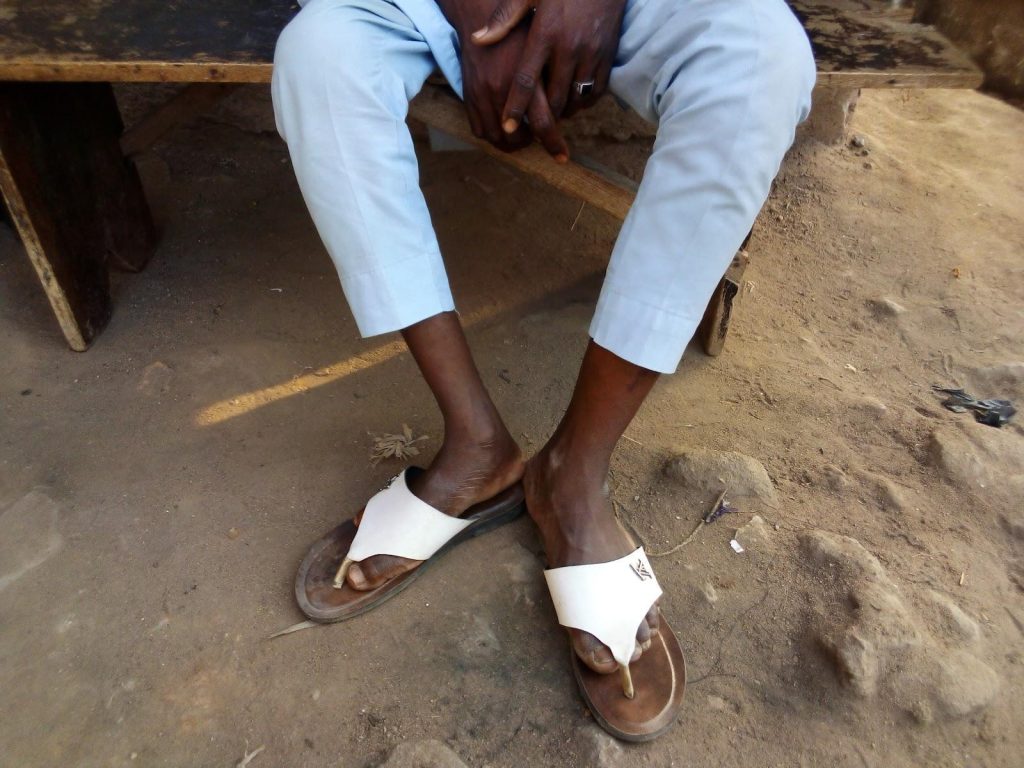 A person wearing white sandals and light blue pants sits on a wooden bench, with crossed arms resting on their lap.