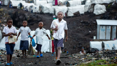 Children walking on a dirt path in front of a refugee camp, carrying items in their hands.