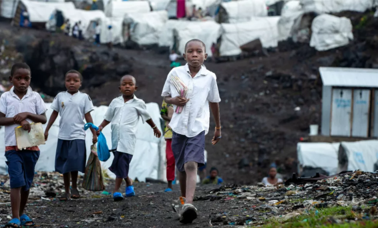 Children walking on a dirt path in front of a refugee camp, carrying items in their hands.